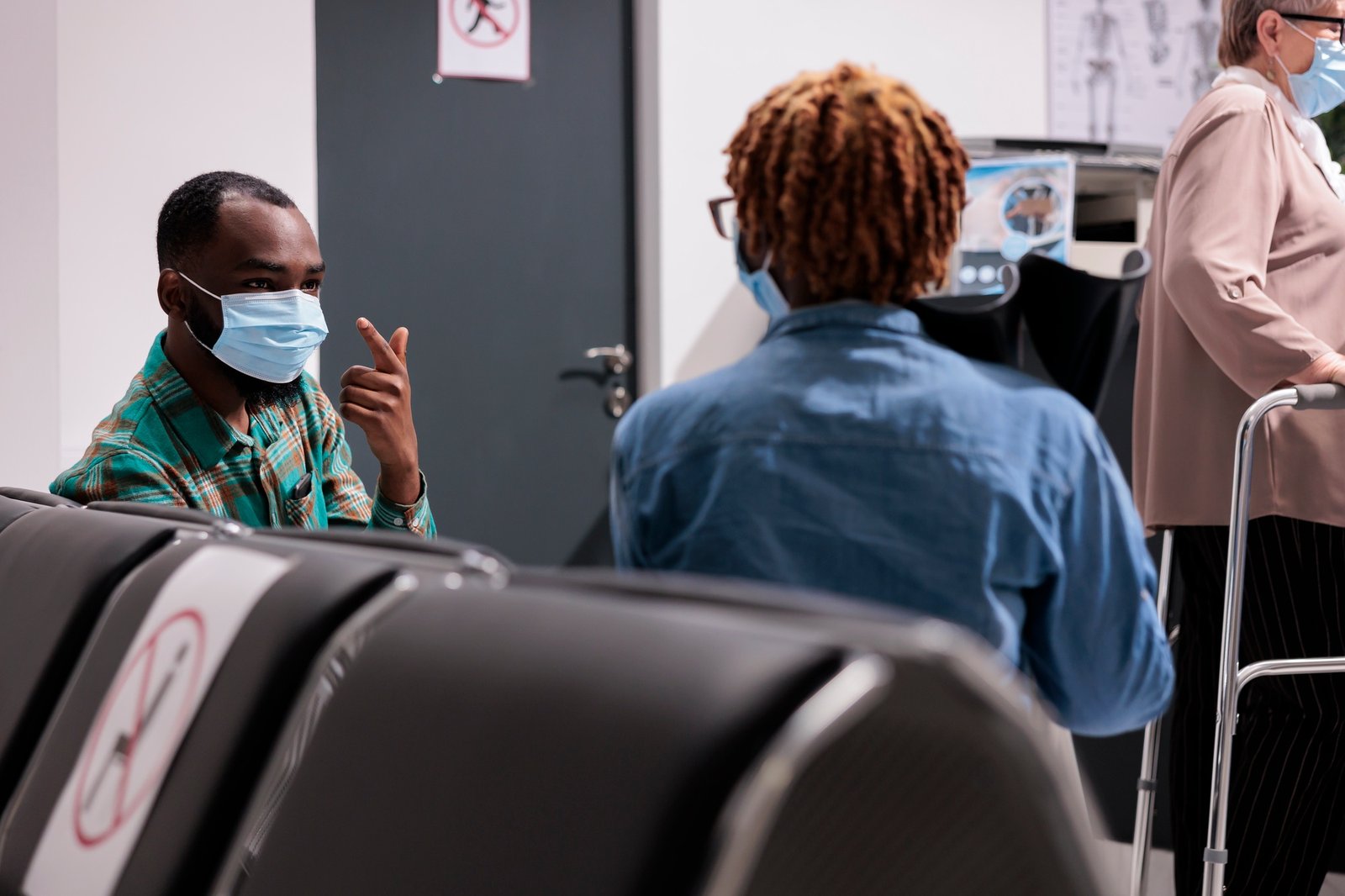 Dos personas con mascarillas conversan en una sala de espera médica; una persona con camisa a cuadros verdes usa lenguaje de señas frente a otra con camisa de mezclilla, mientras al fondo se ve a una persona mayor con andador.