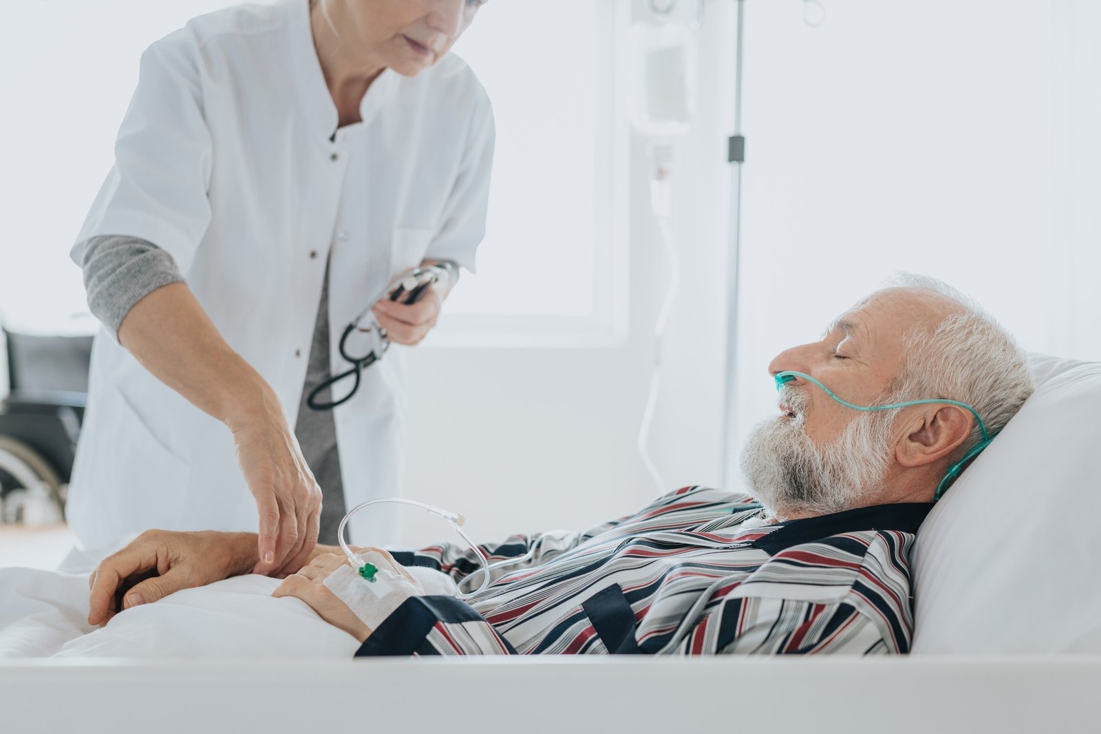 Hombre mayor acostado en una cama de hospital con oxígeno nasal y bata de rayas, atendido por una profesional de salud con bata blanca que le revisa la vía intravenosa.
