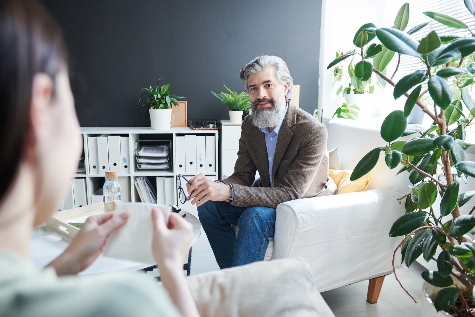 Terapeuta con barba y cabello canoso, sentado en un sillón blanco frente a una mujer; ambos están en una oficina con estanterías y plantas, en un entorno profesional de consulta.